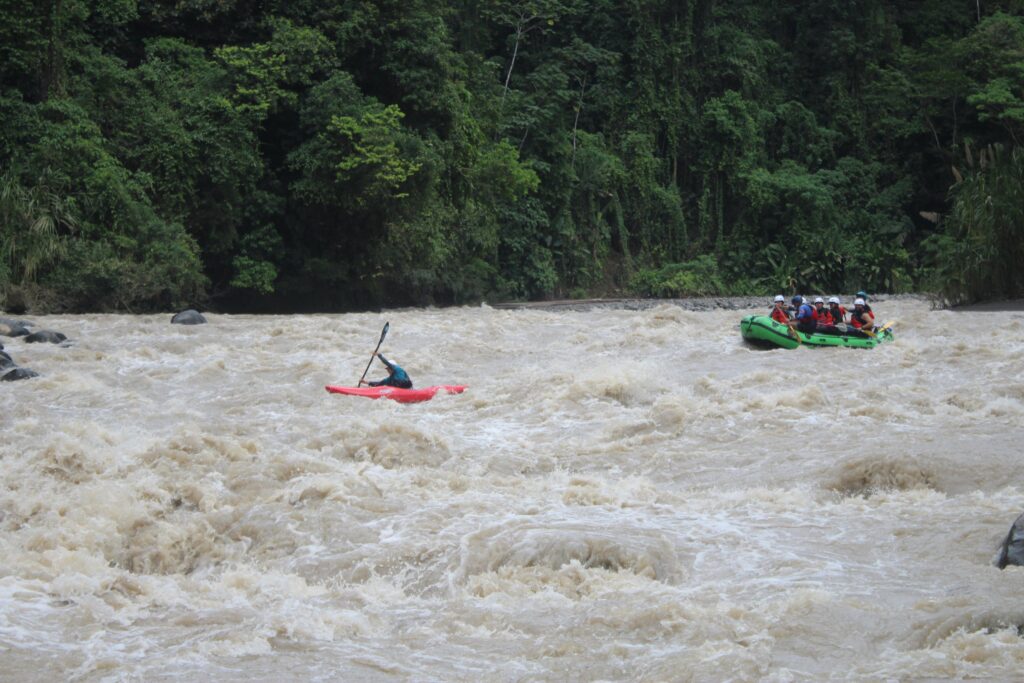 A safety kayaker in a red boat holds position in wide brown rapids as a raft comes down behind