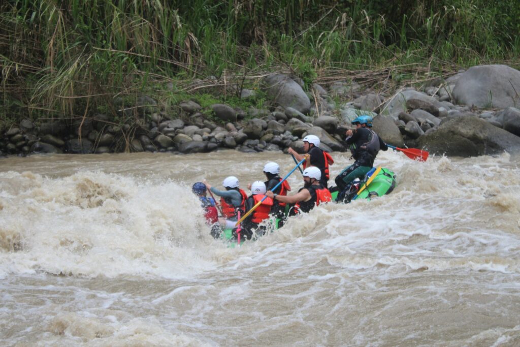 Rafters in a green raft punch through a rapid while their guide at the back calls the strokes