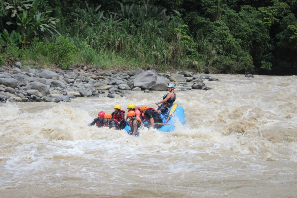 A blue raft of paddlers in orange and red helmets runs a rapid beneath a rocky jungle bank