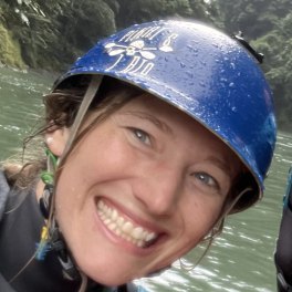 Kayla, smiling, in a blue whitewater helmet on the river